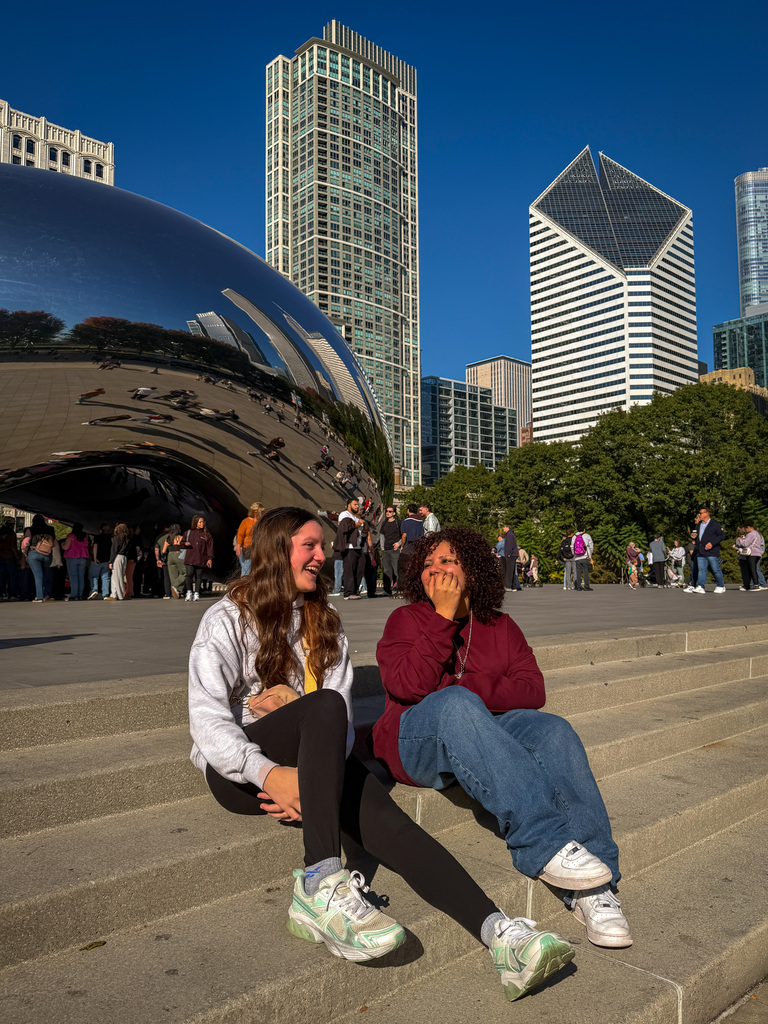 Two students sit on concrete steps in front of a large silver structure. One has a hand covering her mouth.