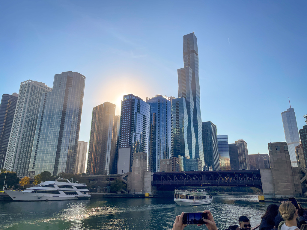 Waterfront view with tall buildings. Boats on water. People taking photos. Sun setting in the distance.