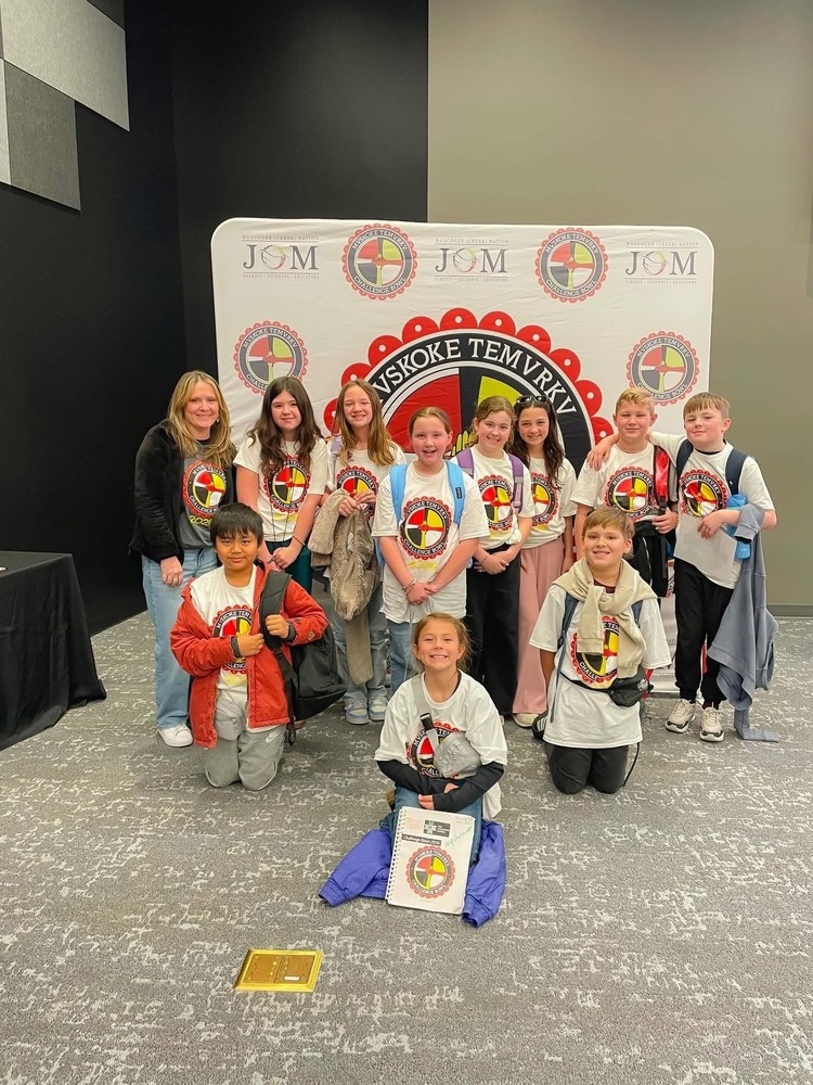 A team of students pose for a photo in front of a banner with their Coach after competing in the Muskogee Creek Nation Challenge Bowl 