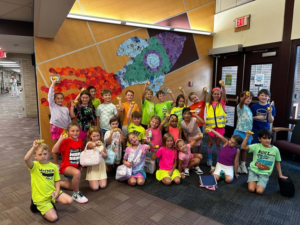 Group of 25 second-graders holding up their yellow rocks in front of the kindness mural