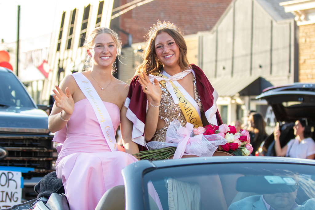 homecoming queen and another girl on the court sitting in a convertible car waving and smiling at the camera