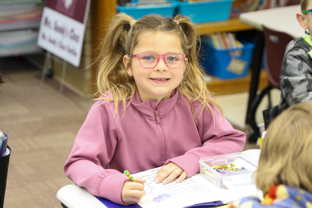 girl wearing glasses coloring and smiling at the camera