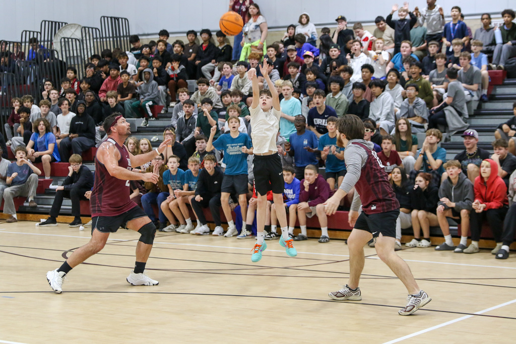 Boy shooting a three pointer mid game with two teachers on each side of him watching the ball in the air. Students behind in the crowd are standing and clapping.