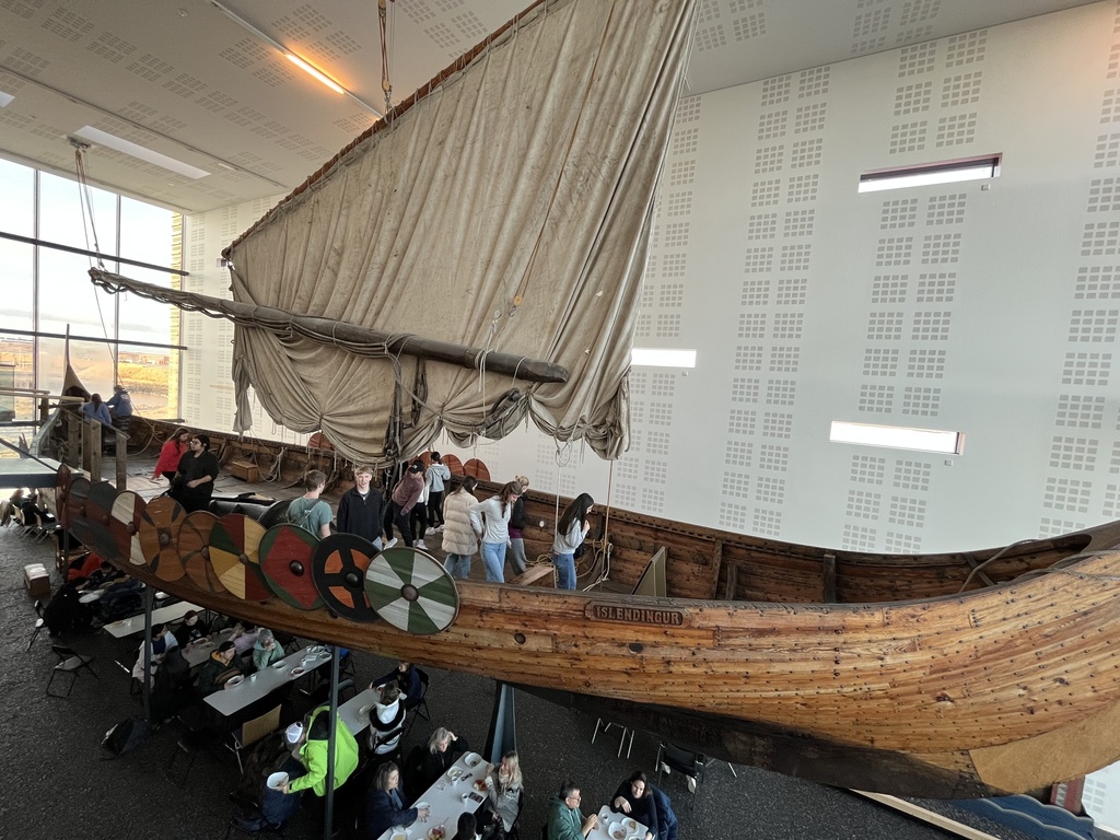 A boat suspended in the air inside a restaurant with students in it. Below the boat are tables with people eating.