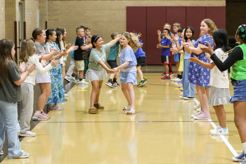 2 girls hand in hand doing a running dance with their peers lined up on the sides clapping and singing