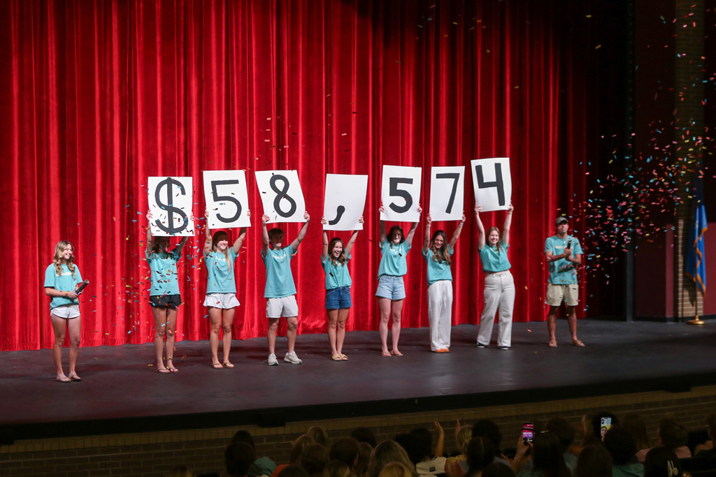 9 JHS STUCO students on the stage. 7 students have signs of the individual numbers of the total. one  students on each side of the total popped confetti into the air. total on the posters they are holding says $58,574