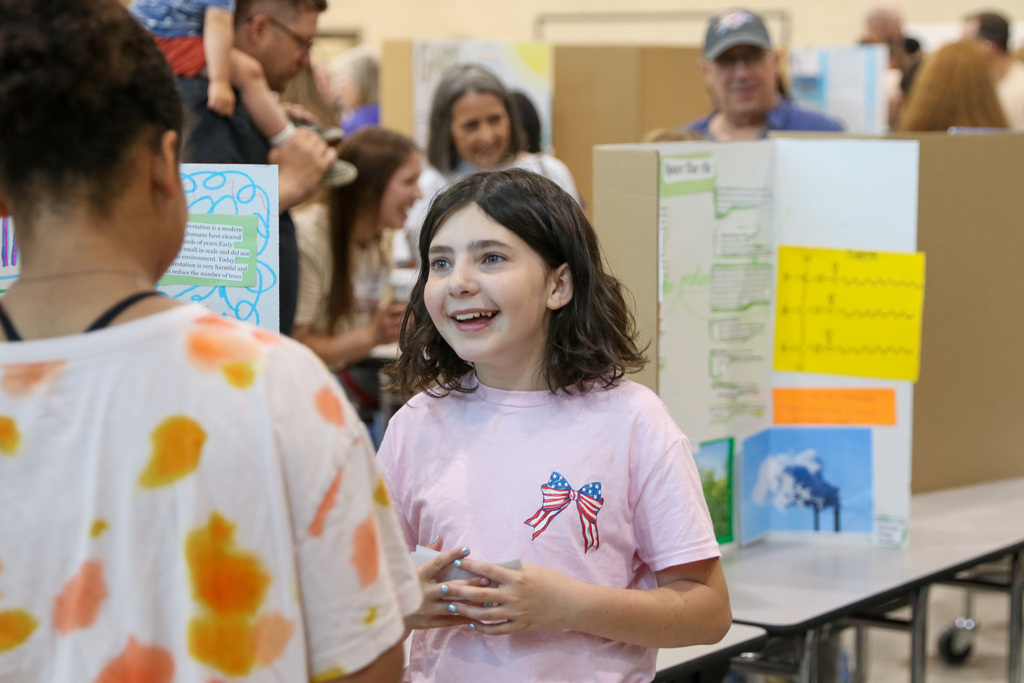 A fourth grade student wearing a pink shirt presents her exhibition project to an adult visitor. She gestures toward a display board while speaking confidently in a classroom setting.