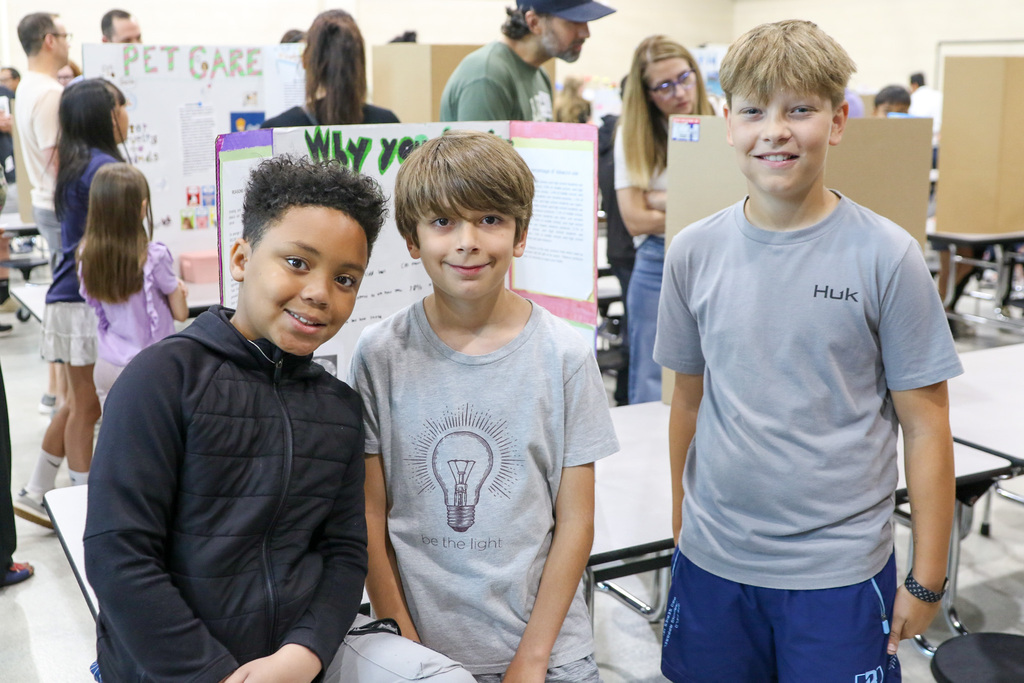 Three fourth grade boys stand together smiling beside their exhibition project display in the classroom. Other students and presentation boards are visible in the background.