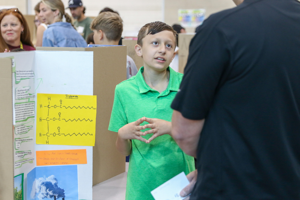 A fourth grade student stands beside his exhibition display while speaking with an adult visitor in the classroom. 