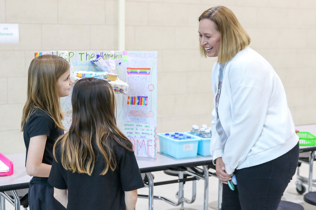Two fourth grade students stand next to their exhibition presentation board while explaining their project to visitors in the classroom.