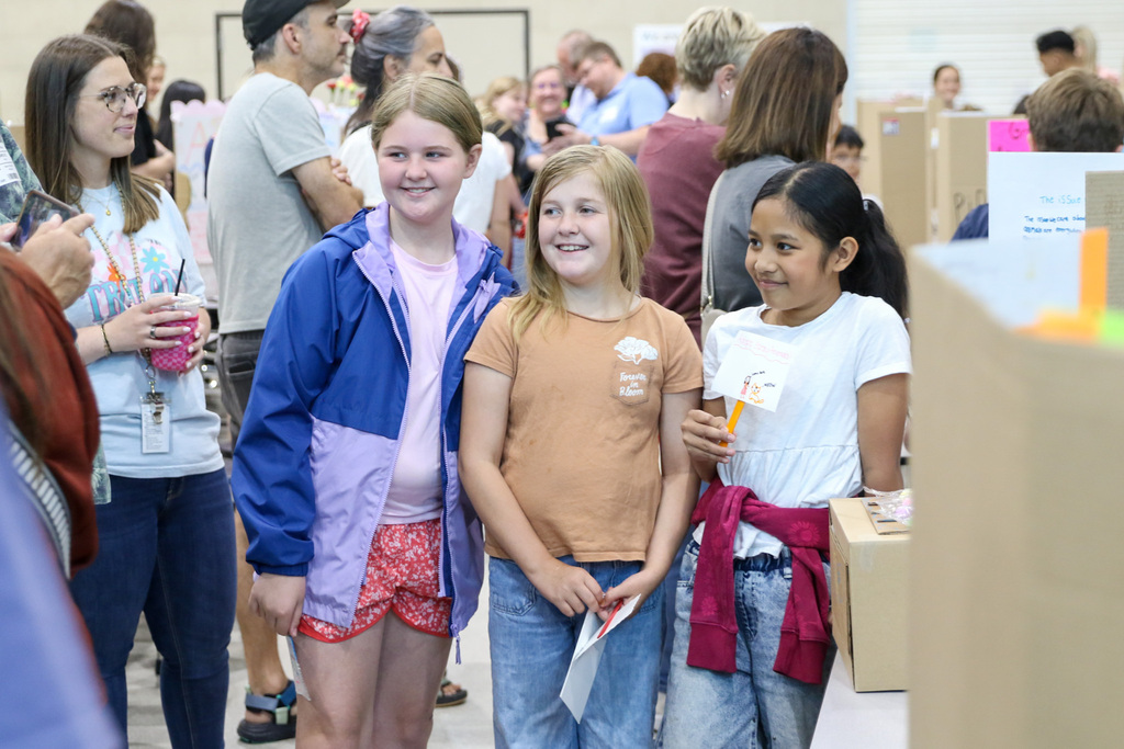 Three fourth grade girls stand together smiling beside their exhibition project display in the classroom. Other students and presentation boards are visible in the background.