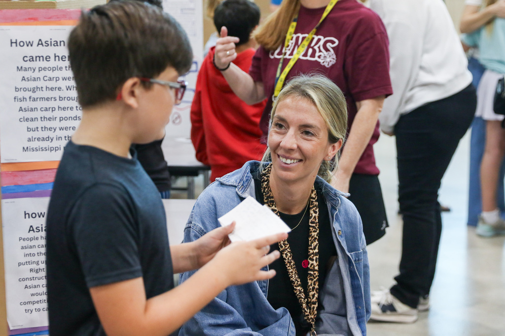 A fourth grade student presents to  an adult visitor while sitting beside an exhibition project display. Other students and presentation boards are visible in the background.
