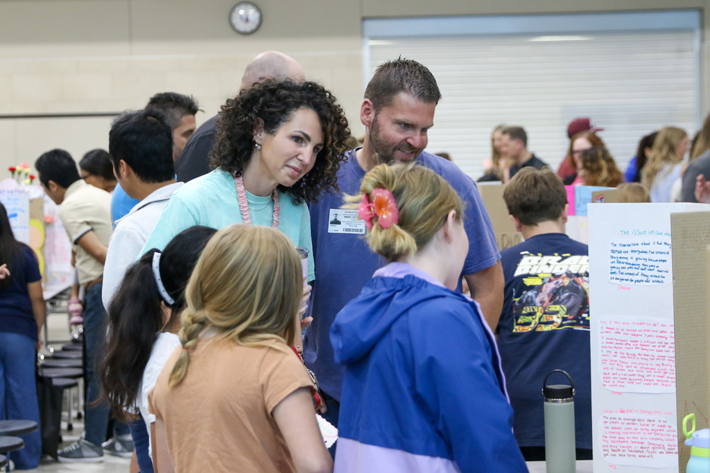 3 fourth grade students stand beside their exhibition display while speaking with visitors stand at a table. One student points toward information on the project board during the discussion.