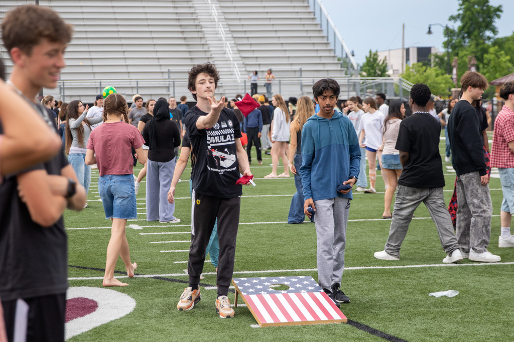 Two students stand on a football field playing cornhole beside an American flag-themed game board while other students gather and walk around in the background.