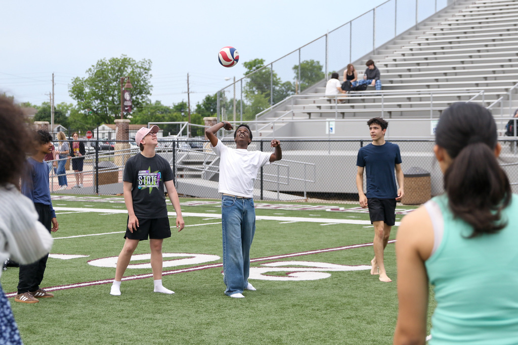 Several students spread out across a football field participate in an outdoor activity. Two students stand in the center with arms raised to hit a volleyball while others watch nearby, with stadium bleachers in the background.
