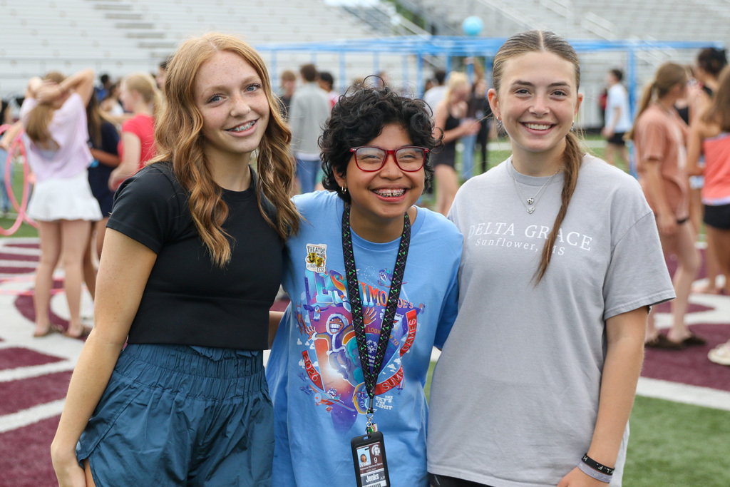 Three students stand together smiling on a football field during an outdoor school event. The student in the middle wears a blue shirt and a lanyard, while the two students beside them pose with their arms around each other.