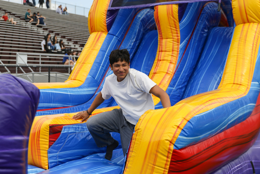 A student in a white short-sleeve shirt slides down a colorful inflatable obstacle course with bright blue, yellow, and orange sides. Stadium bleachers are visible in the background.