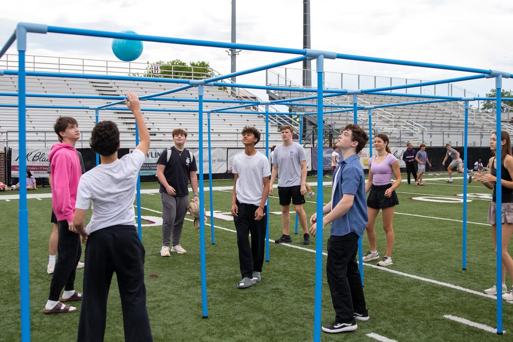 Students stand around a blue metal frame structure on a football field, appearing to participate in a game or activity while other students mingle in the background.