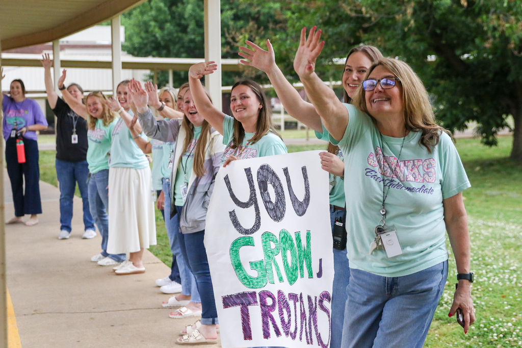 teachers standing outside waving to students with a sign that says you grow, trojans!