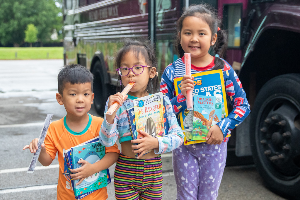 three children with popsicles in one hand and books in the other smiling for a photo infront of the bookmobile