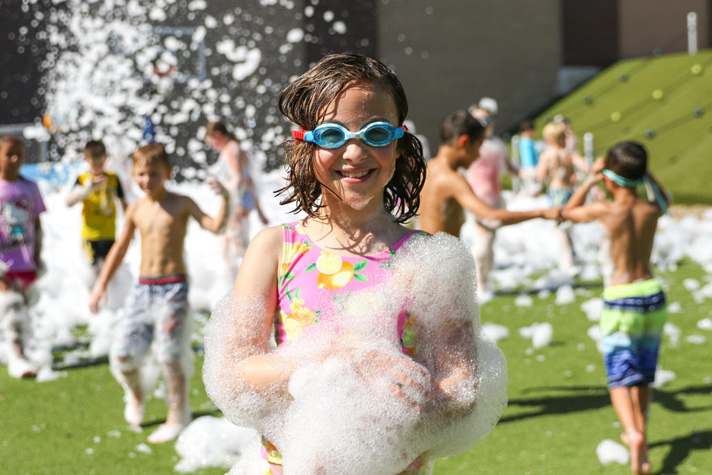 girl covered in foam with swim goggles on smiles for a photo with other kids playing in the foam behind her