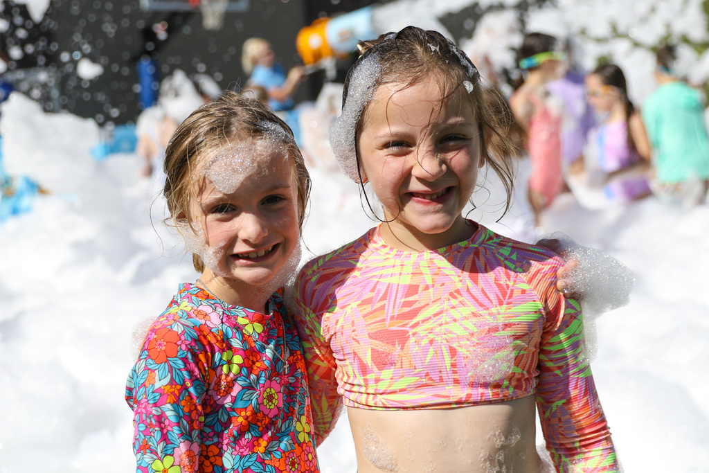 two girls smiling for the photo with bubbles on their head. foam and kids playing are in the background.