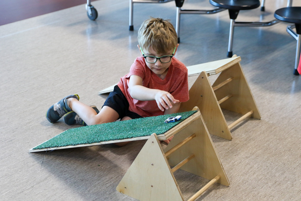 boy sitting on the ground next to a wooden ramp that has turf on it rolling a toy car down the ramp