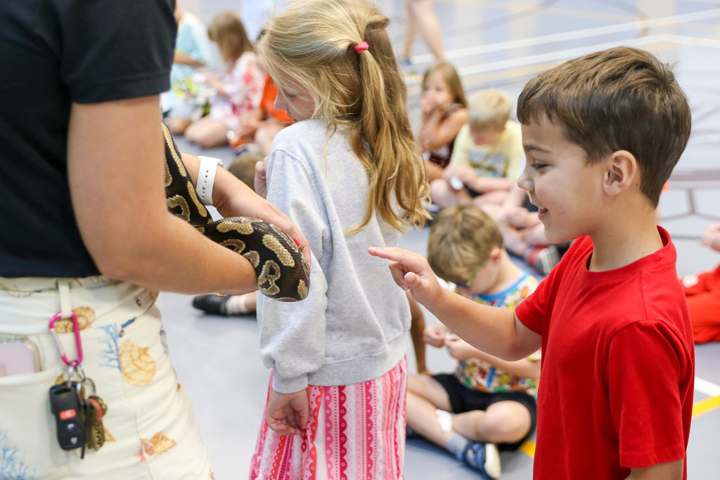 boy smiling with his finger out to pet a snake that is wrapped around the instructors arm.