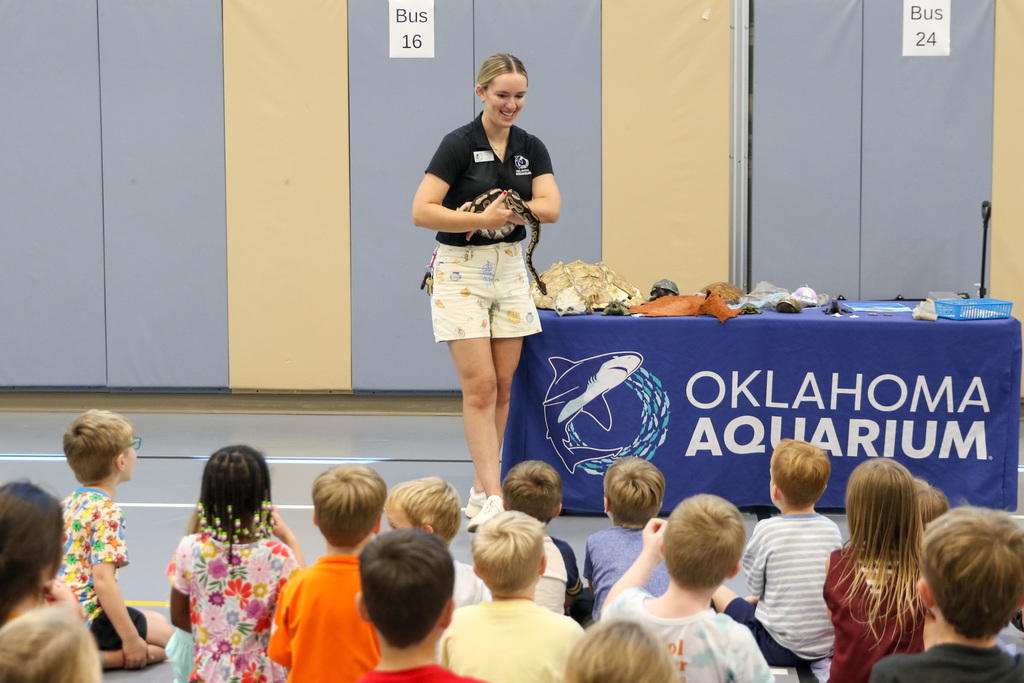Instructor with a snake on her arm smiling down at the students. table next to her with turtle shells and reptile bones on it. table cloth says oklahoma aquarium