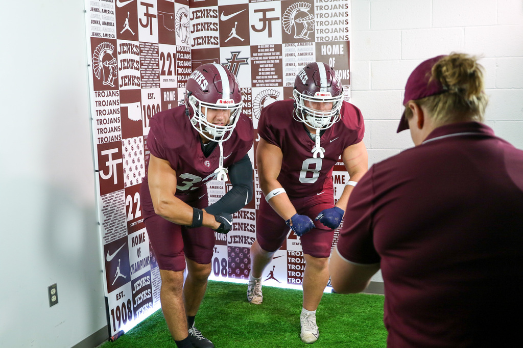 two football players leaning towards the camera flexing. they are in front of a backdrop that is collage of jenks trojans athletics.