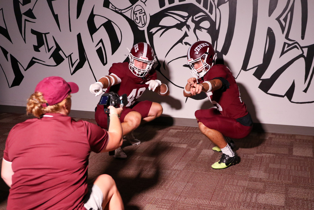 two football players squted down pointing at the camera in front of a graffiti that says jenks trojans with a trojan head in the middle of it.