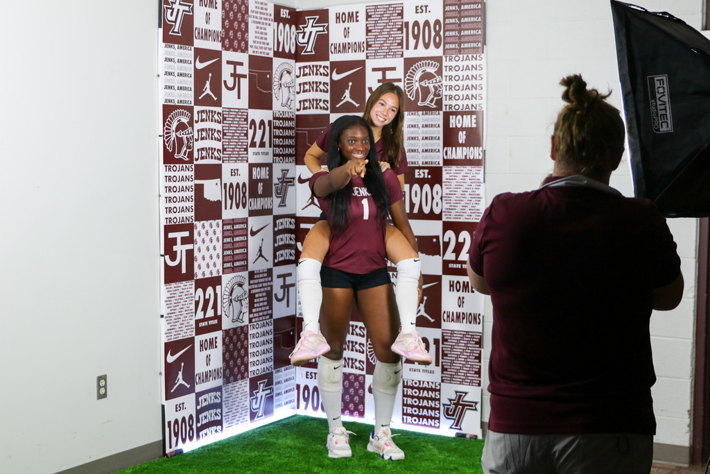 volleyball player standing and pointing to the camera with a teammate on her back smiling. they are in front of a backdrop that is collage of jenks trojans athletics.