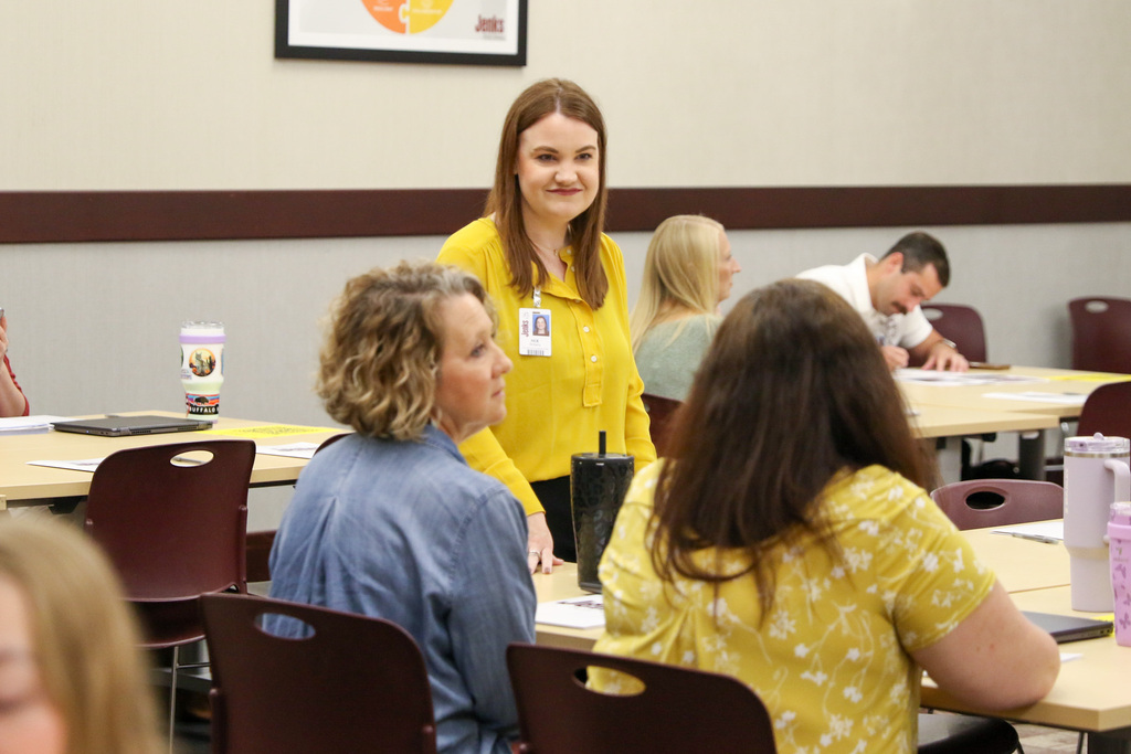 staff standing and smiling while listening to a new teacher who is sat at a table