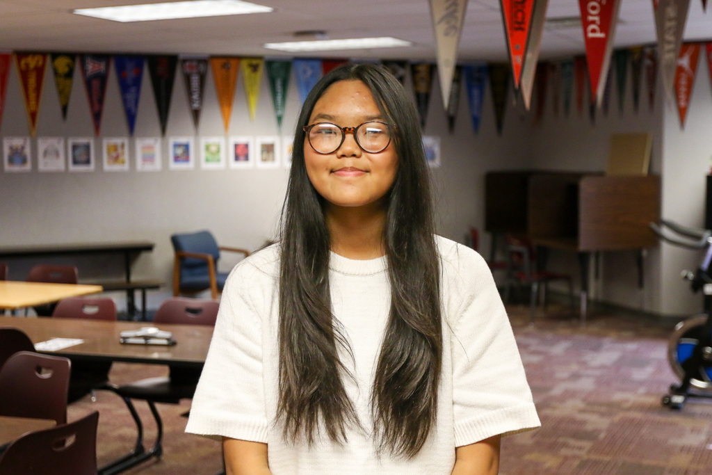8th grader young scholar Vung Buansing smiling for a photo with college flags on the walls behind her