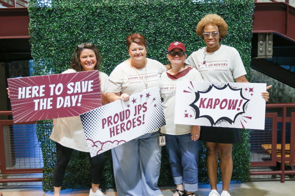 4 transportation staff members infront of a greenery wall with signs that say here to save the day, proud jps heroes, and kapow!