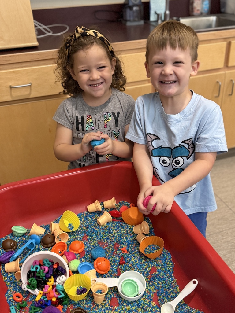 Two smiling young children play together with colorful scoops, cups, and toys in a sensory bin filled with rainbow-colored rice.