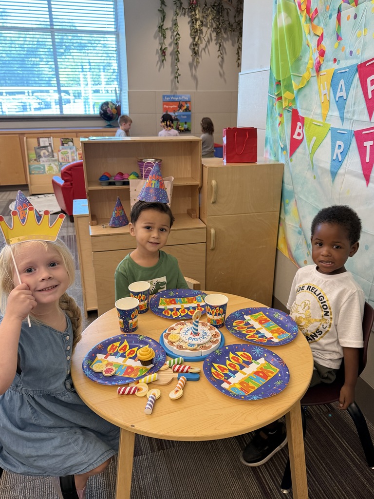 Three young children smile around a small table decorated for a birthday celebration, wearing party hats and holding a birthday crown.