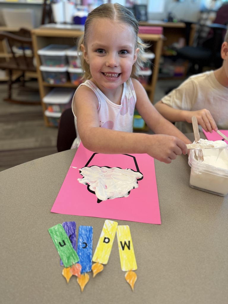 A smiling young girl spreads white fluffy paint onto a pink paper craft at a classroom table while colorful craft pieces sit nearby.
