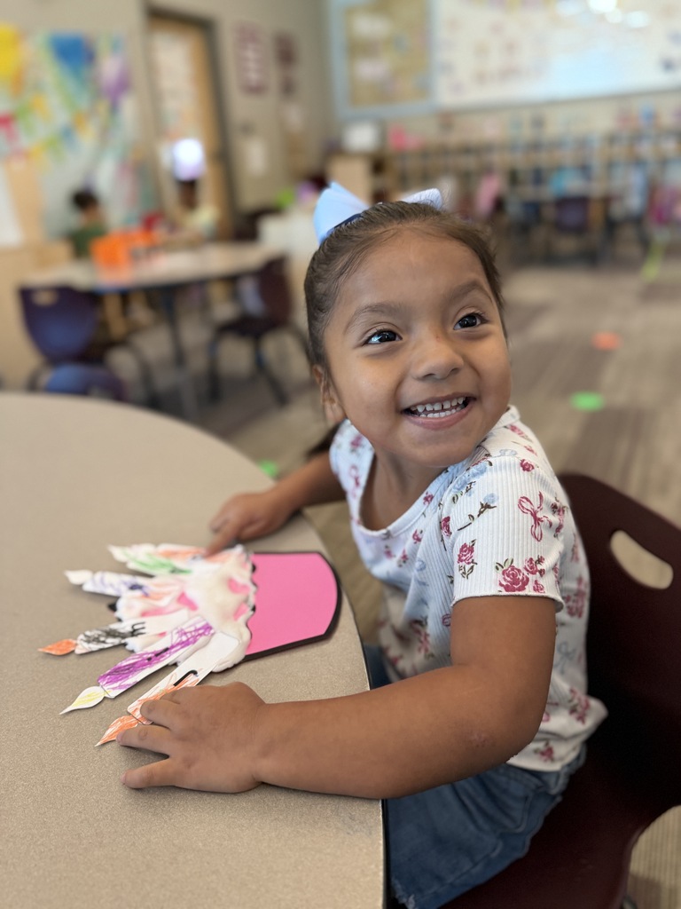 A smiling young girl wearing a blue bow works on a colorful classroom craft at a table.