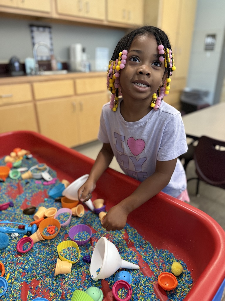 A young girl with pink and yellow beaded braids plays with colorful scoops and toys in a sensory bin filled with rainbow-colored rice.