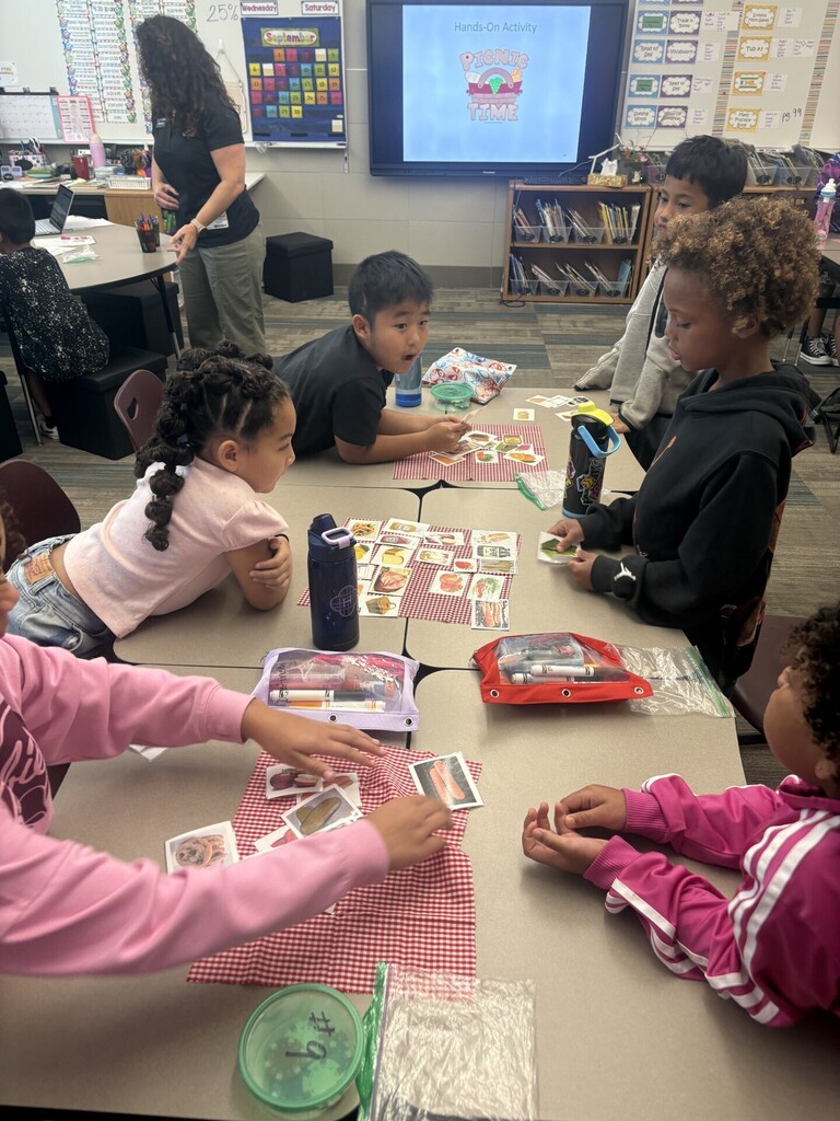 Students work together at classroom tables during a hands-on “Picnic Time” activity, sorting and arranging picture cards on red-and-white checkered mats while their teacher assists.