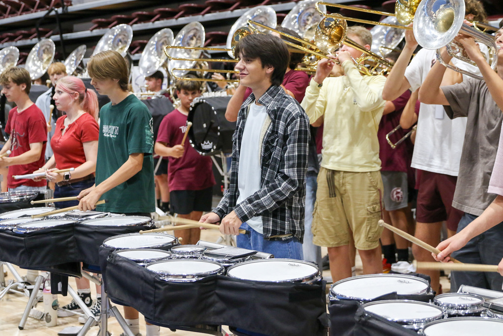 up close photo of drumline drumming with students behind with trombones playing