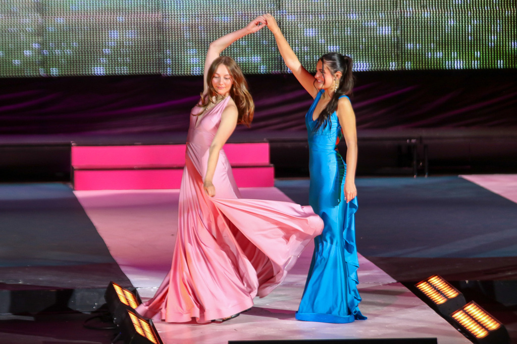 two girls in prom dresses. they have an arm up and one is twirling the other. the one being twirled has her dress flying in the wind