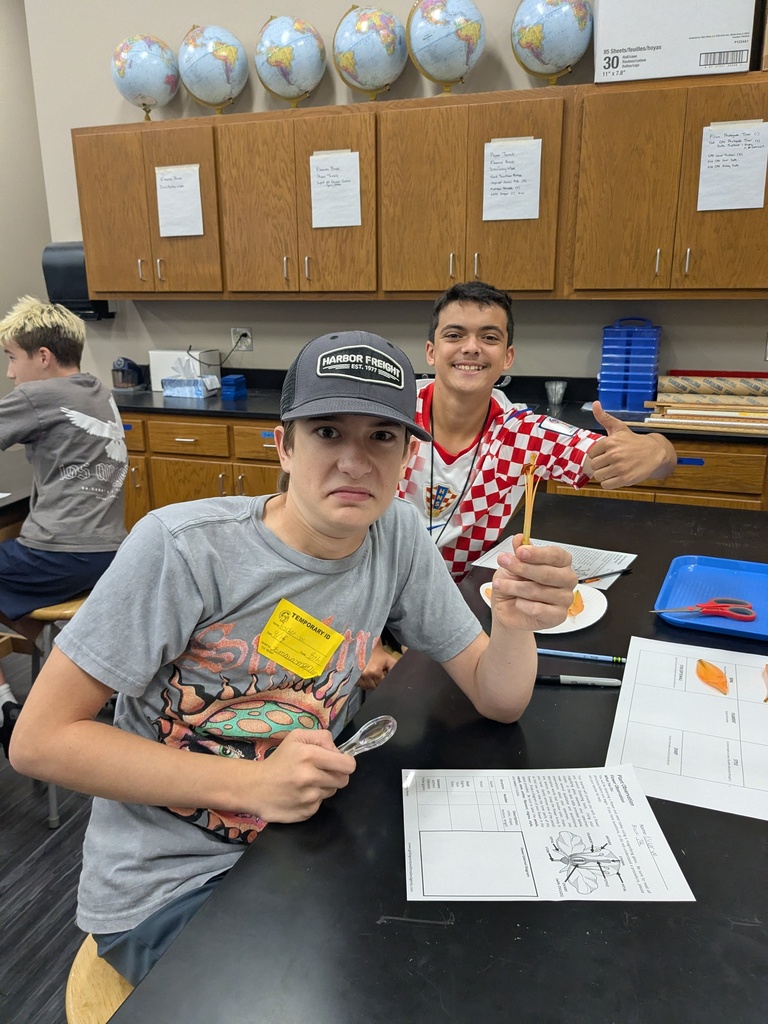 student holding flower in foreground with student in background giving thumbs up