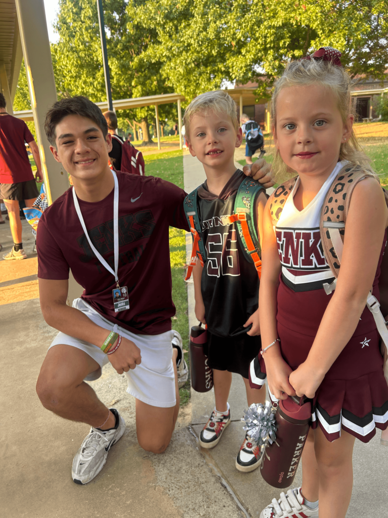 A highschool student kneels next to two young elementary students all wearing Jenks themed clothing.