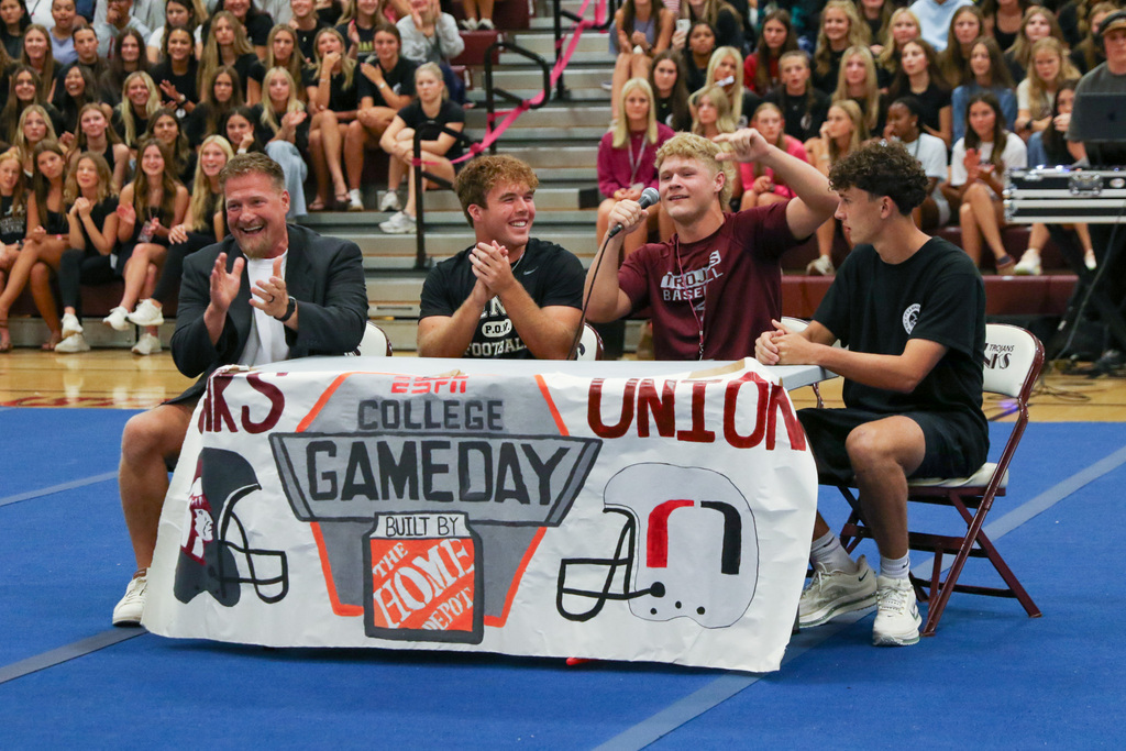 table with a banner on it that says ESPN college game day with a jenks football helment and a union football helmet. behind the table sits a couch and two football players and another student