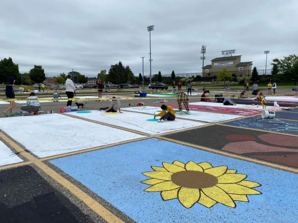 Students painting parking spots. Most spots are white, one is blue with a yellow and brown sunflower, another is black with red geometric shapes.