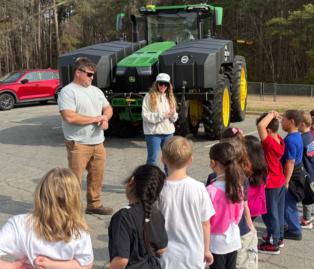 Image of Jared and Caroline Barefoot speaking to first graders about their career in farming. A tractor is behind them as they are speaking.
