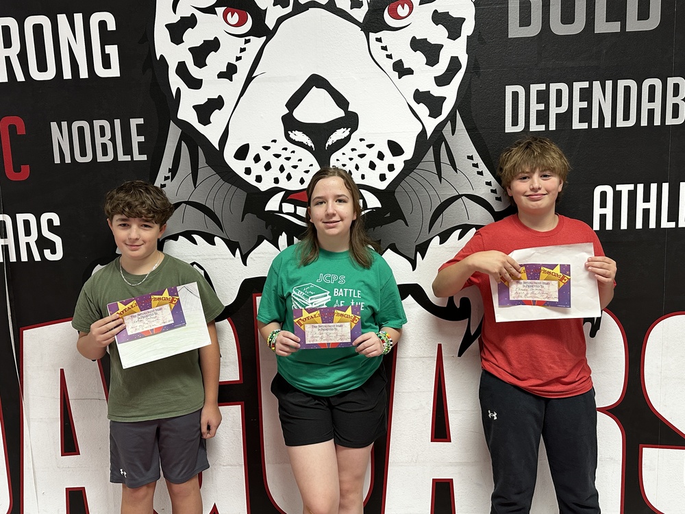 3 students standing in front of the jaguar mural holding certificates 