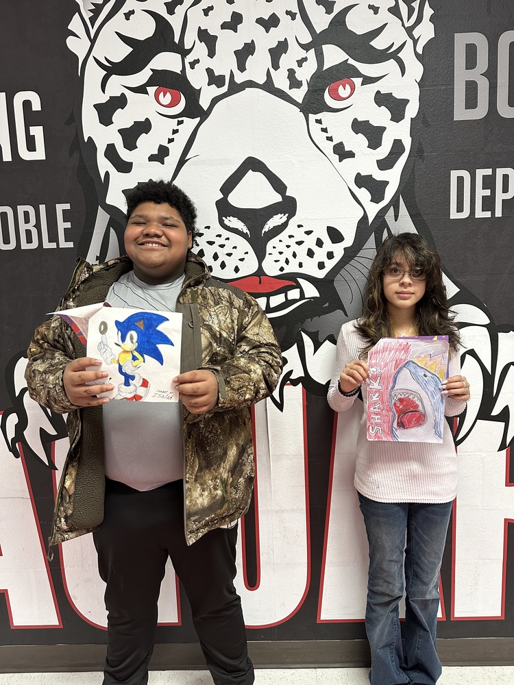 Picture of a male and female NJMS students holding artwork in front of the jaguar mural 